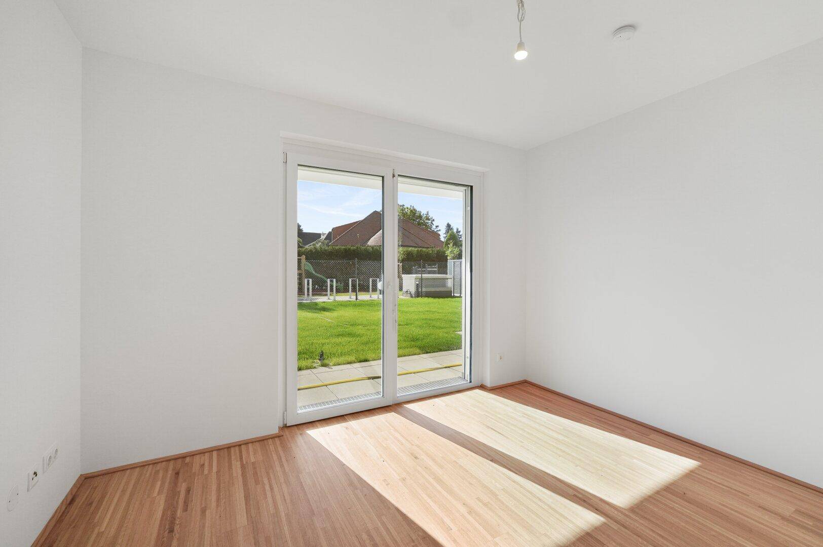 Geräumiges Zimmer mit Holzboden und großen Fenstern, die viel Tageslicht hereinlassen und Gartenblick bieten.