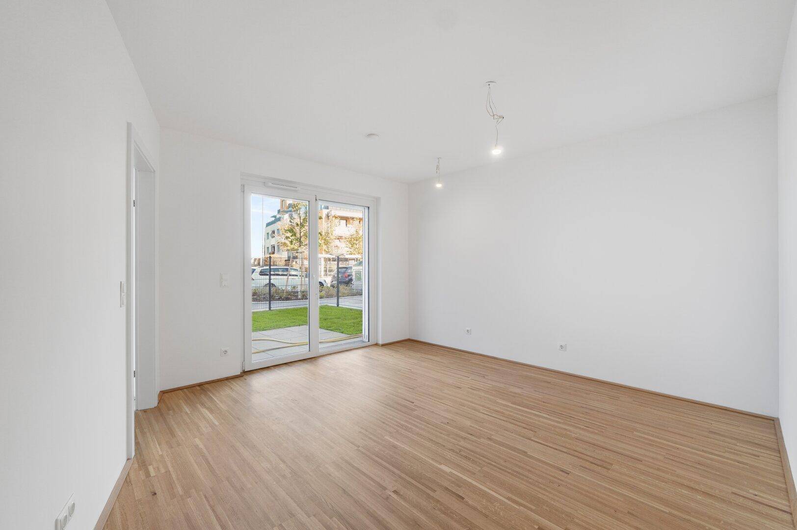 Geräumiges Wohnzimmer mit hellem Holzboden und Zugang zu einem Außenbereich durch eine große Glastür.