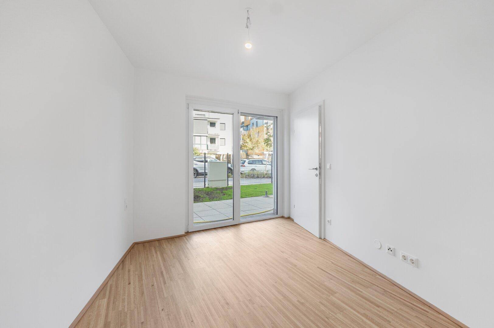 Helles Zimmer mit Holzboden und großer Schiebetür, die Zugang zu einem kleinen Gartenbereich bietet.