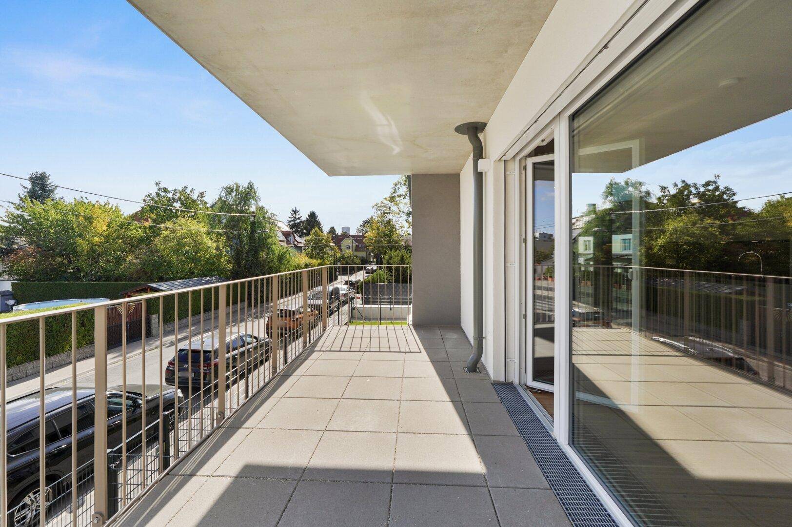 Geräumiger Balkon mit Fliesenboden und Metallgeländer, bietet Blick auf die Straße und Bäume.