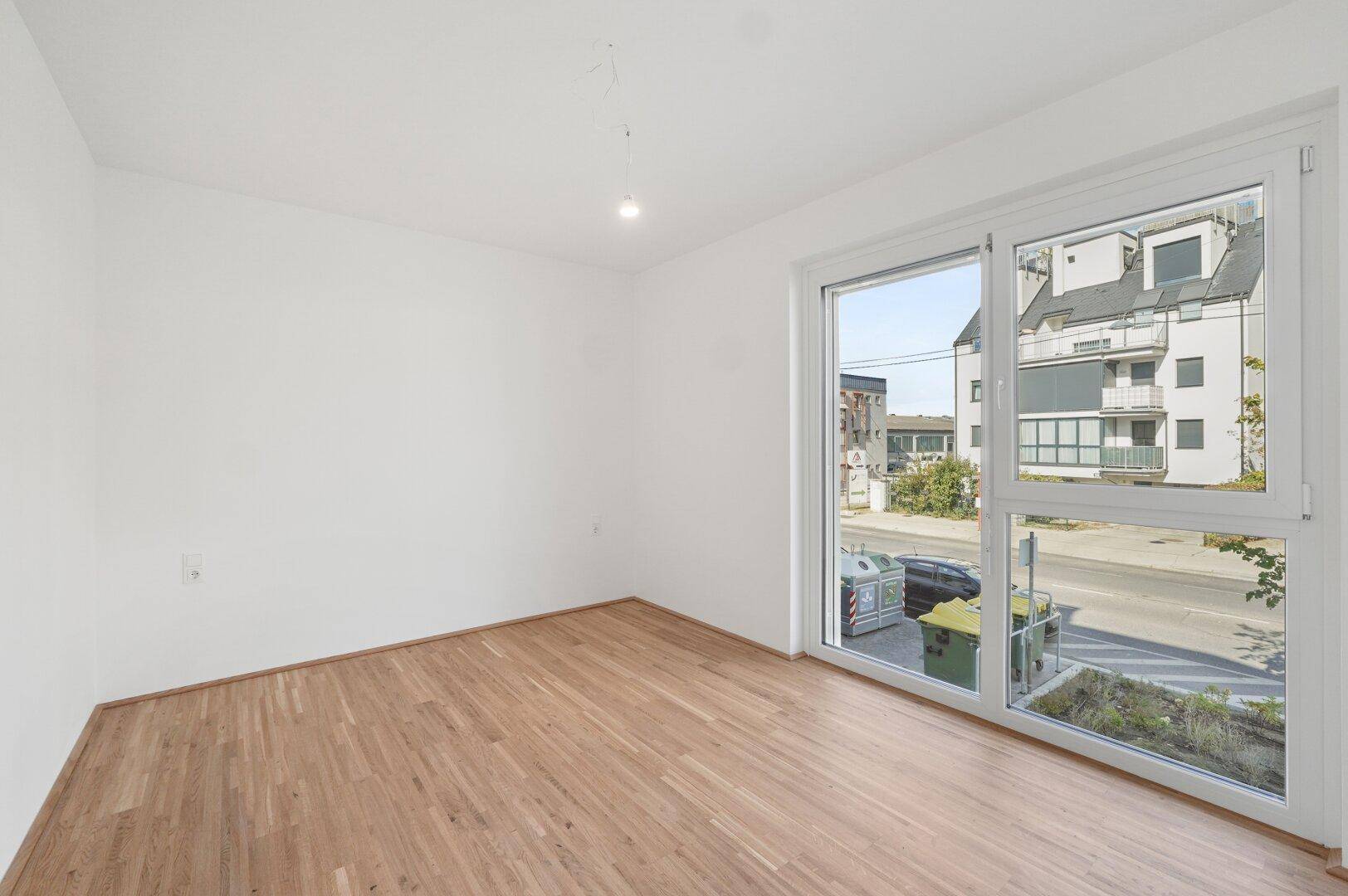 Unmöbliertes Schlafzimmer mit großen Fenstern und Blick auf die Straße und umliegende Gebäude.