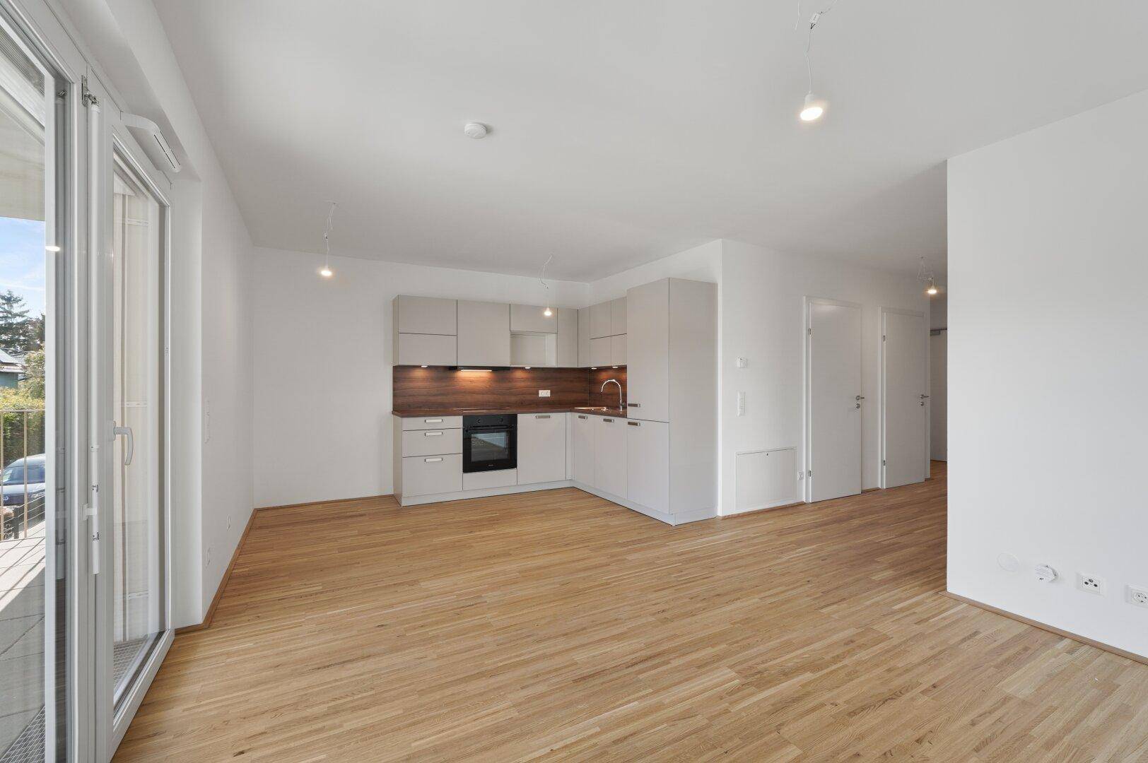 Geräumiger Wohnbereich mit integrierter Küchenzeile und hellem Holzfußboden in Neubauwohnung.