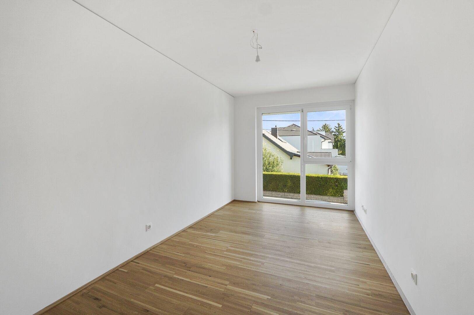 Helles Schlafzimmer mit Parkettboden und großem Fenster mit Blick auf die Hecke.