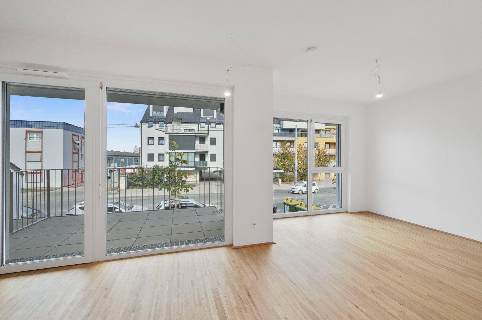 Geräumiger Wohnbereich mit großen Fenstern, Holzboden und Zugang zu einem breiten Balkon.
