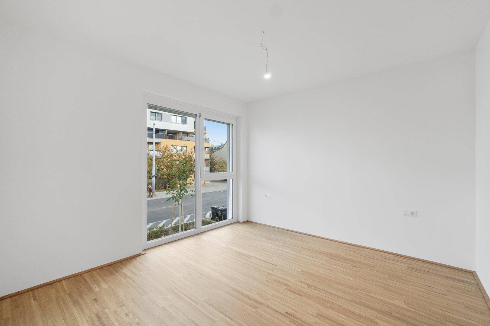 Leerer Raum mit hellem Parkettboden und großem Fenster mit Blick auf die Straße.