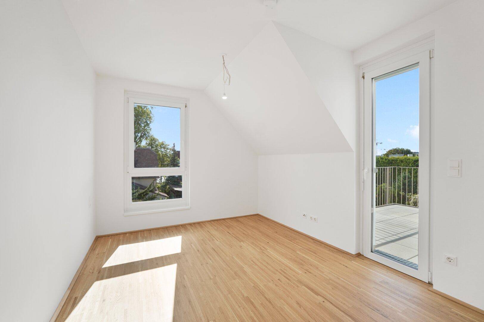 Lichtdurchflutetes Zimmer mit Holzboden, Dachschräge und Zugang zu einem Balkon.