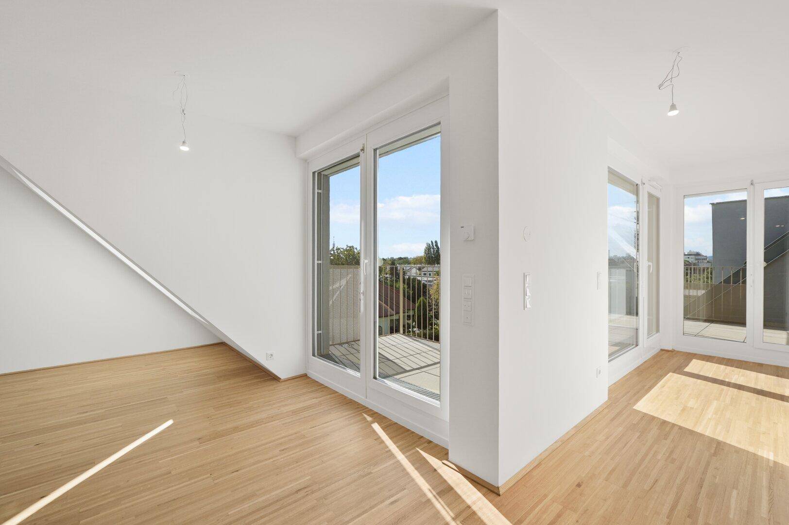 Lichtdurchfluteter Wohnbereich mit Parkettboden und großen Fenstern, die auf einen Balkon führen.