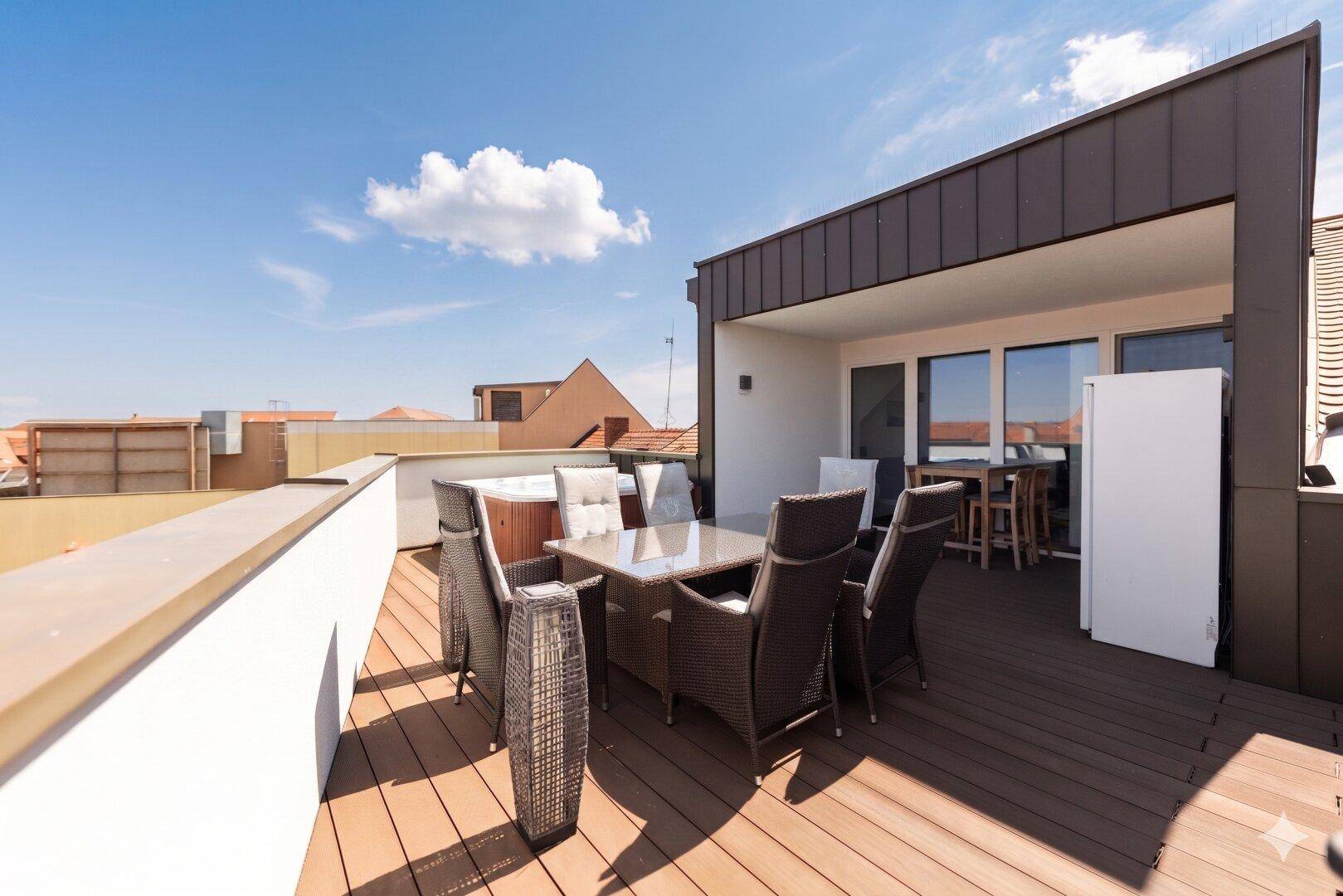 Geräumige Dachterrasse mit Holzboden, gemütlichen Loungemöbeln und einem Whirlpool unter blauem Himmel.