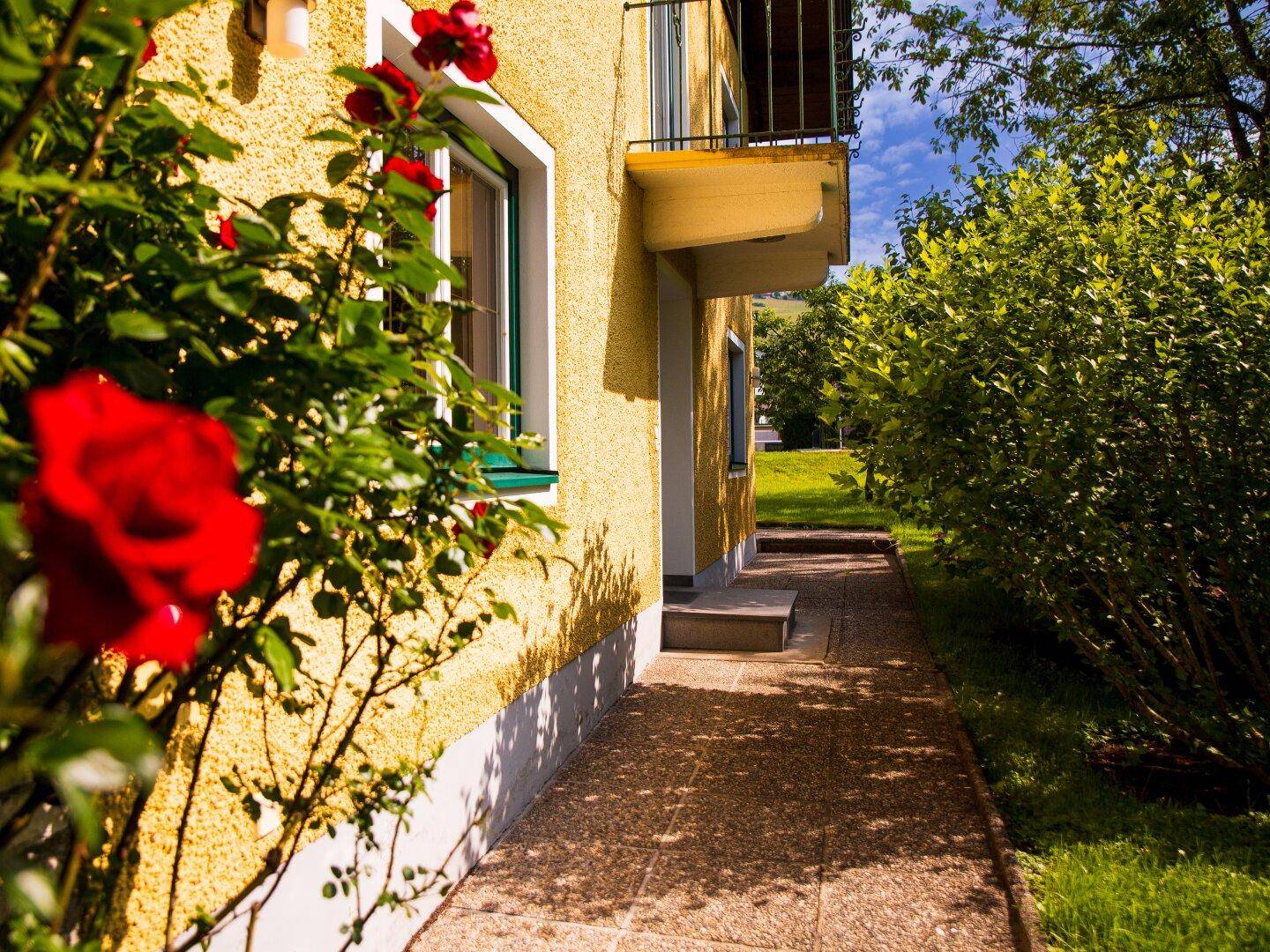 Gelbe Hausfassade mit kleinem Balkon, gepflastertem Weg und blühenden Rosenbüschen im Vordergrund.