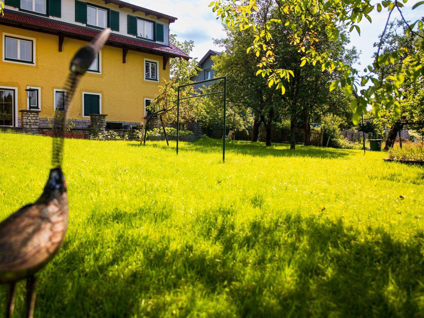 Großer, sonniger Garten mit weitläufiger Rasenfläche, Bäumen und einem gelben Haus im Hintergrund.