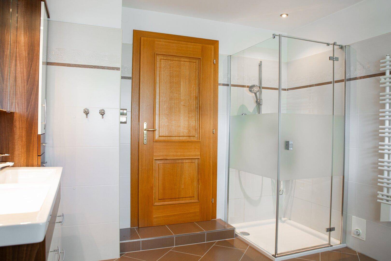 Modernes Badezimmer mit Glasdusche, Holztür und beheiztem Handtuchhalter in hellen Farbtönen.