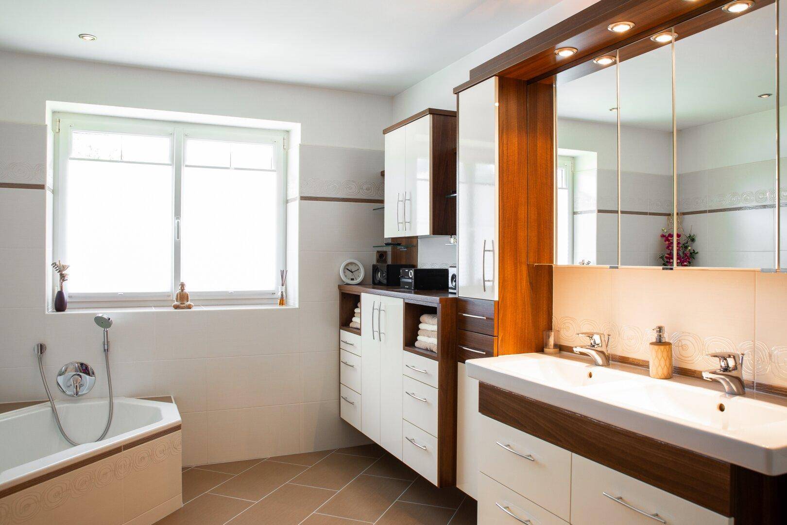 Helles Badezimmer mit Eckbadewanne, Doppelwaschbecken und großem Spiegelschrank unter einem Fenster.