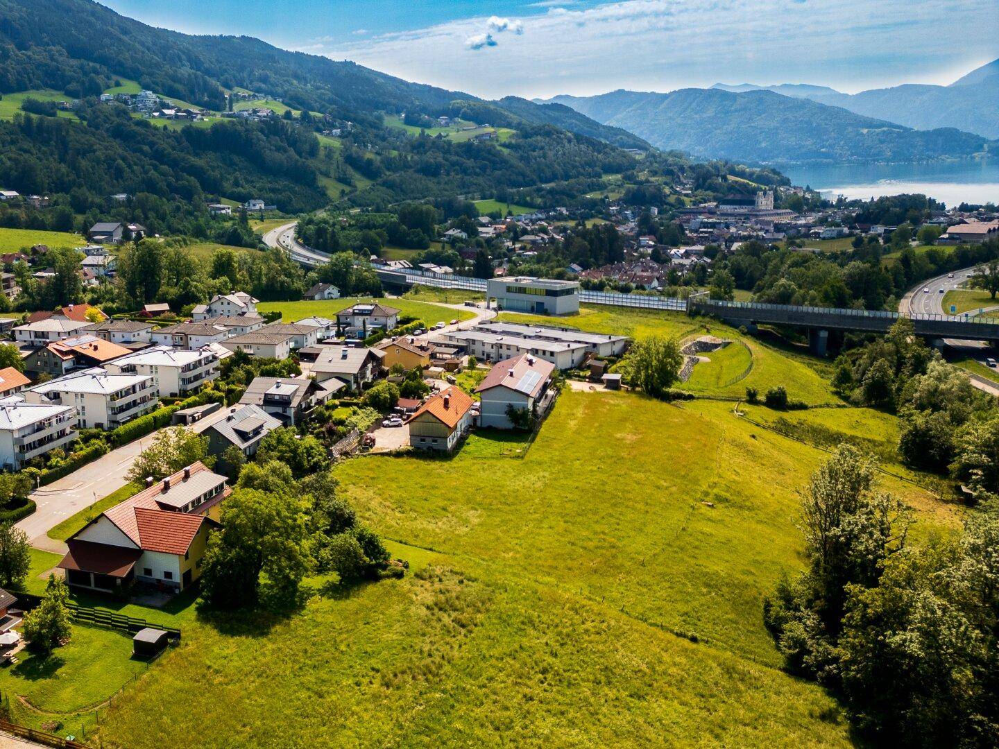 Panoramische Luftaufnahme einer Siedlung in einem Tal, umgeben von grünen Hügeln, Bergen und einem See.