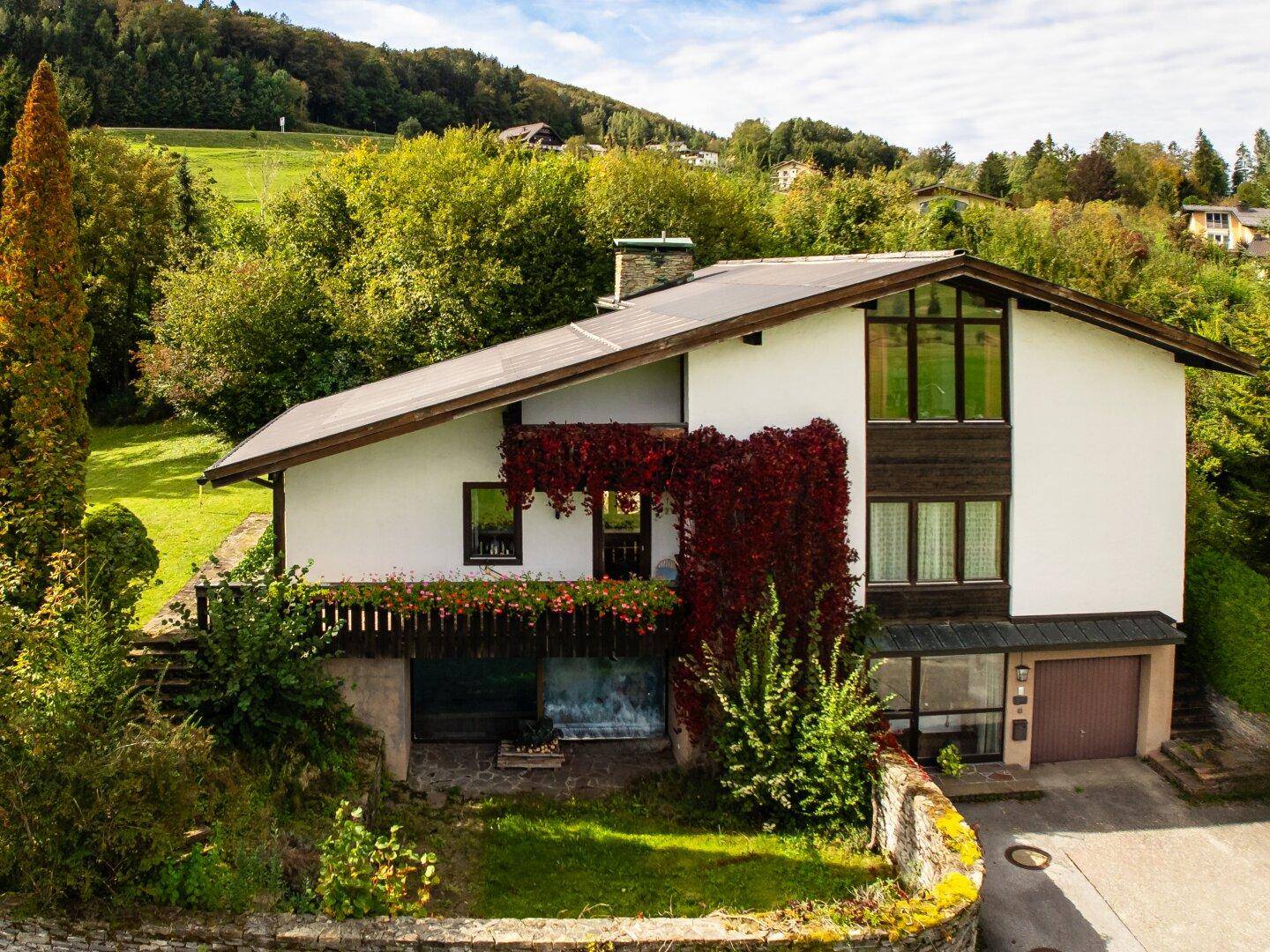 Ansicht eines Hauses mit Balkon, Garage und üppiger Vegetation an einem sonnigen Hang.