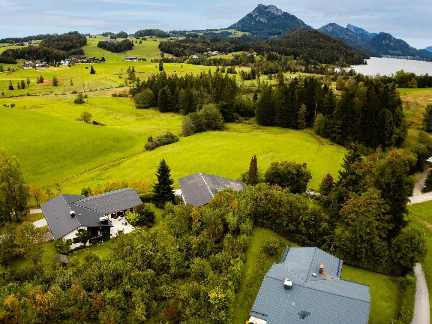 Luftaufnahme einer Wohnsiedlung in einer hügeligen Landschaft mit See und Bergen im Hintergrund.