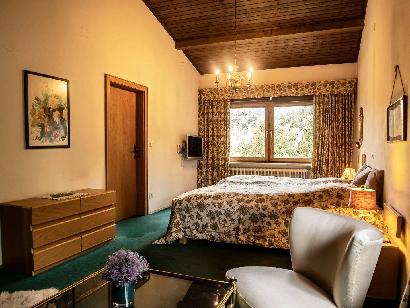 Schlafzimmer mit Doppelbett, Holzbalkendecke, grünem Teppich und traditionellen Möbeln.