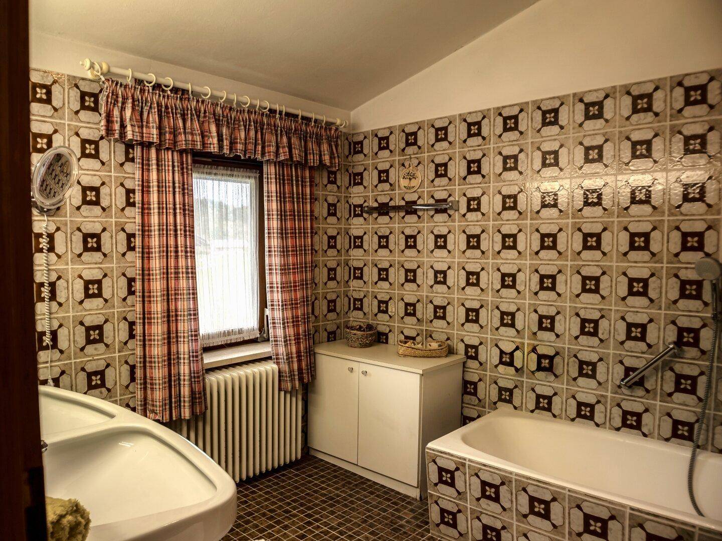 Badezimmer mit gemusterten Fliesen, Badewanne, Waschbecken und einem Fenster mit karierten Vorhängen.