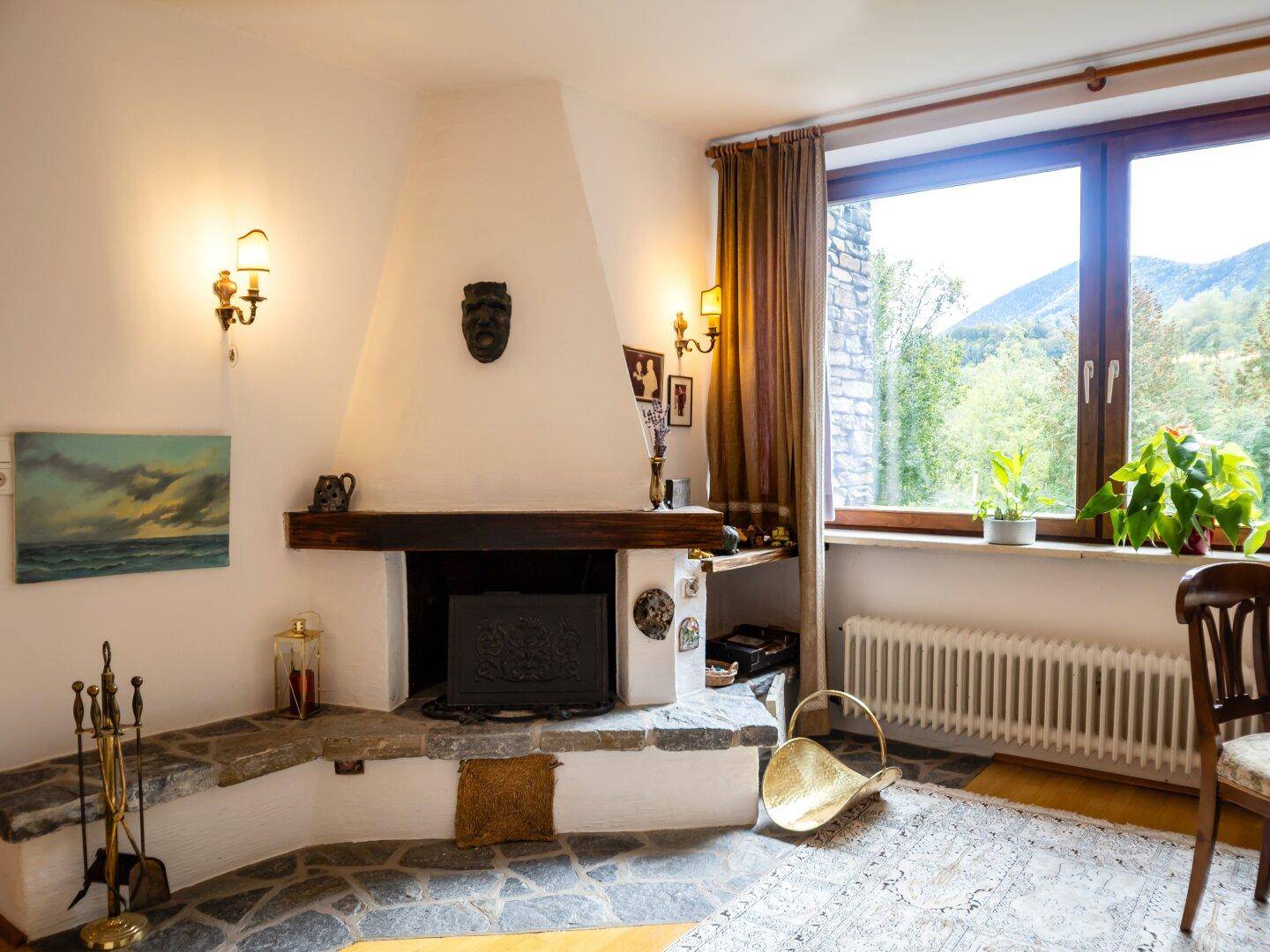 Wohnzimmer mit großem Steinkamin, Holzbalken und einem Fenster mit Blick ins Grüne.