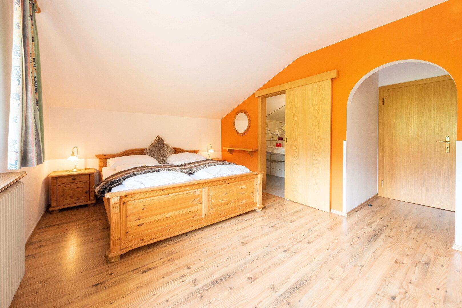 Helles Schlafzimmer im Dachgeschoss mit Holzboden, orangefarbener Wand und Zugang zum Badezimmer.