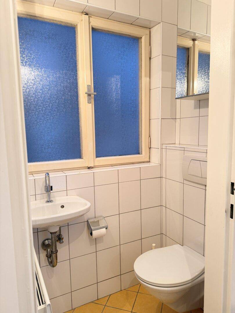 Kleines Gäste-WC mit weißen Fliesen, wandhängendem WC, Waschbecken und einem Fenster mit Milchglas.
