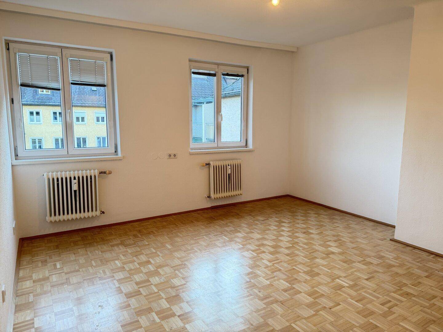 Helles, leeres Zimmer mit Fischgrätparkettboden, weißen Wänden und zwei Fenstern mit Heizkörpern.
