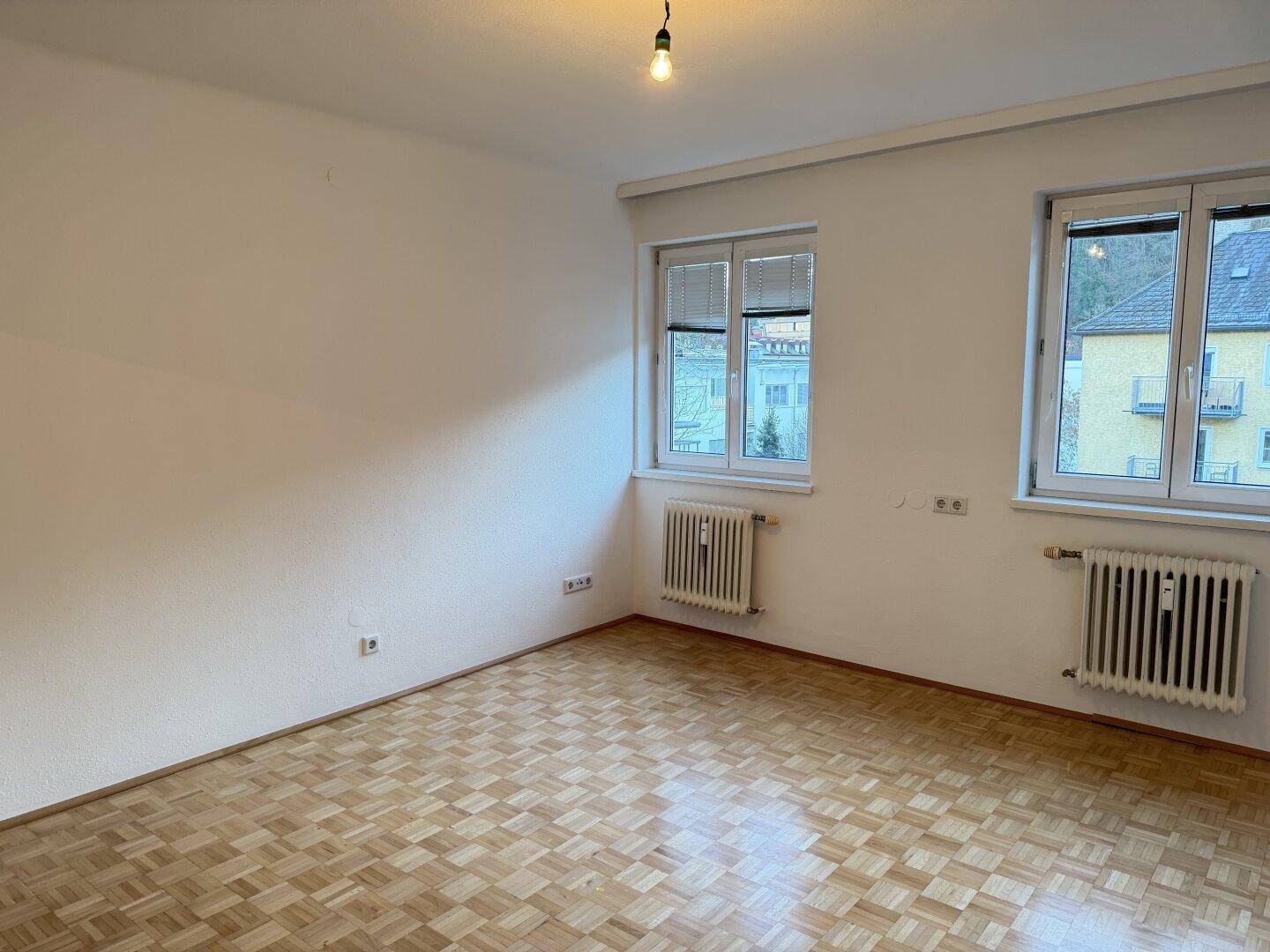 Geräumiges, unmöbliertes Zimmer mit Parkettboden und zwei großen Fenstern, die viel Licht hereinlassen.