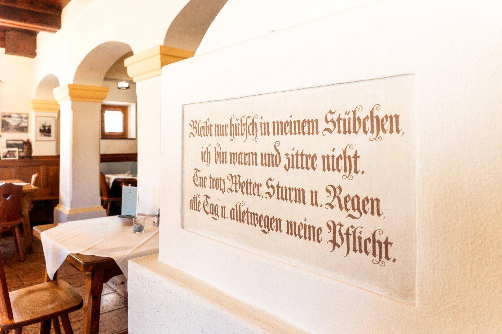 Gemütlicher Essbereich mit traditionellem Kachelofen, Holzbänken und einem Spruch an der Wand.