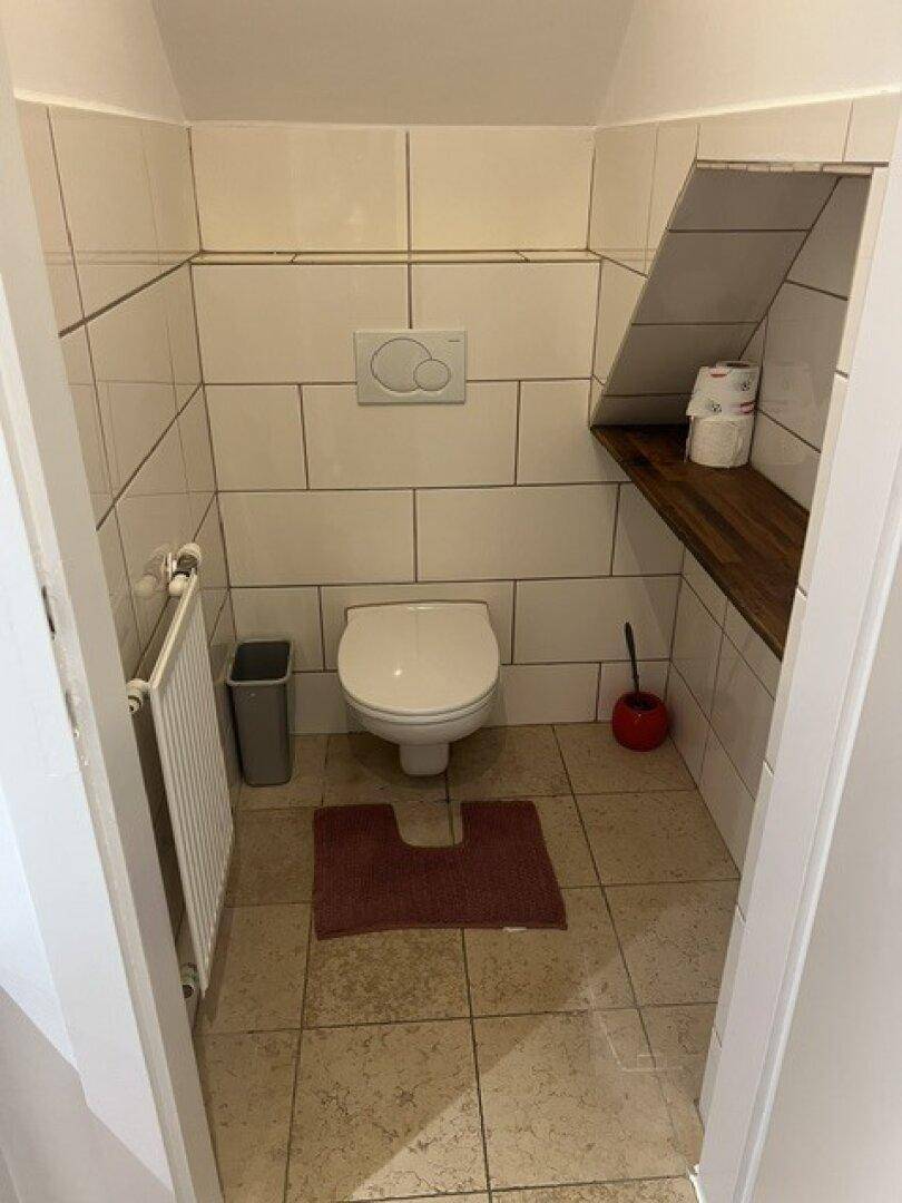 Separates WC mit weißen Fliesen, wandhängender Toilette und praktischer Ablagefläche.