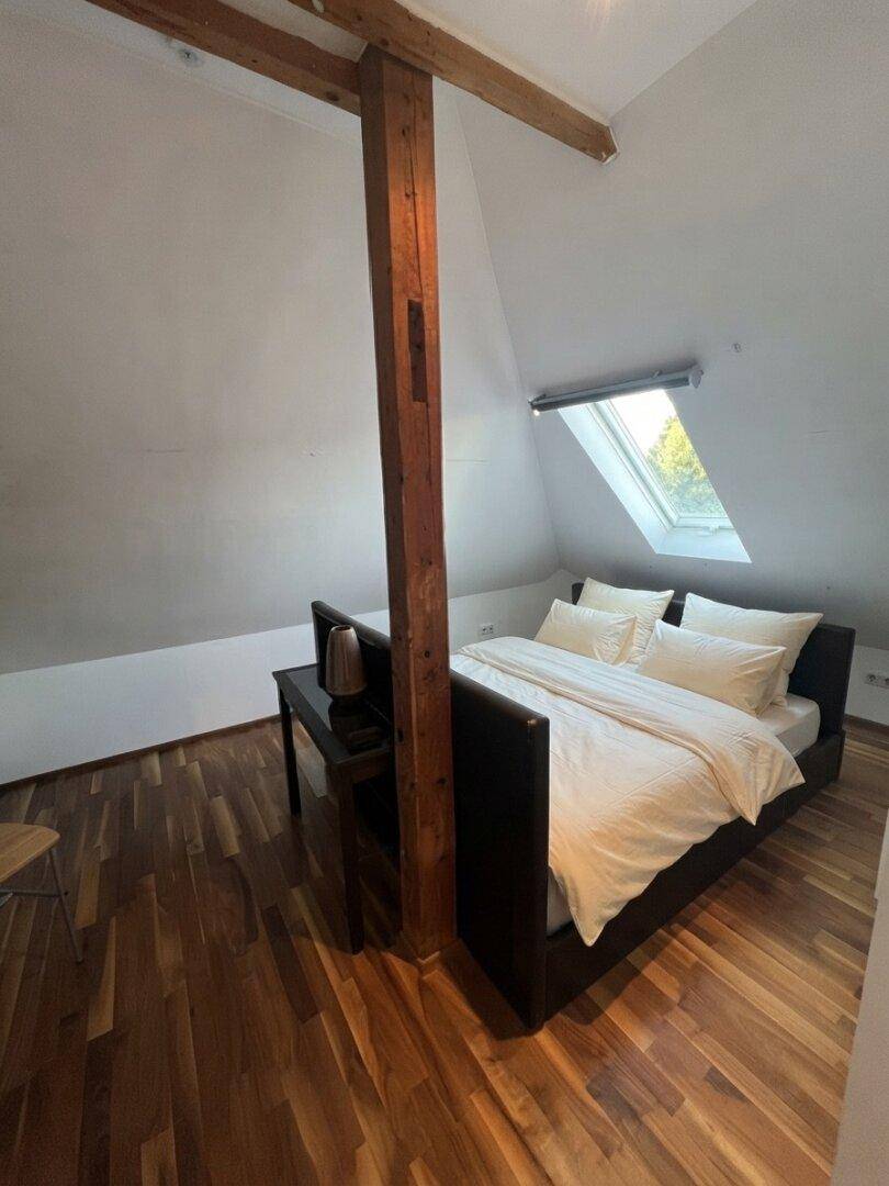 Gemütliches Schlafzimmer im Dachgeschoss mit Holzbalken, Holzboden und Dachfenster für natürliches Licht.