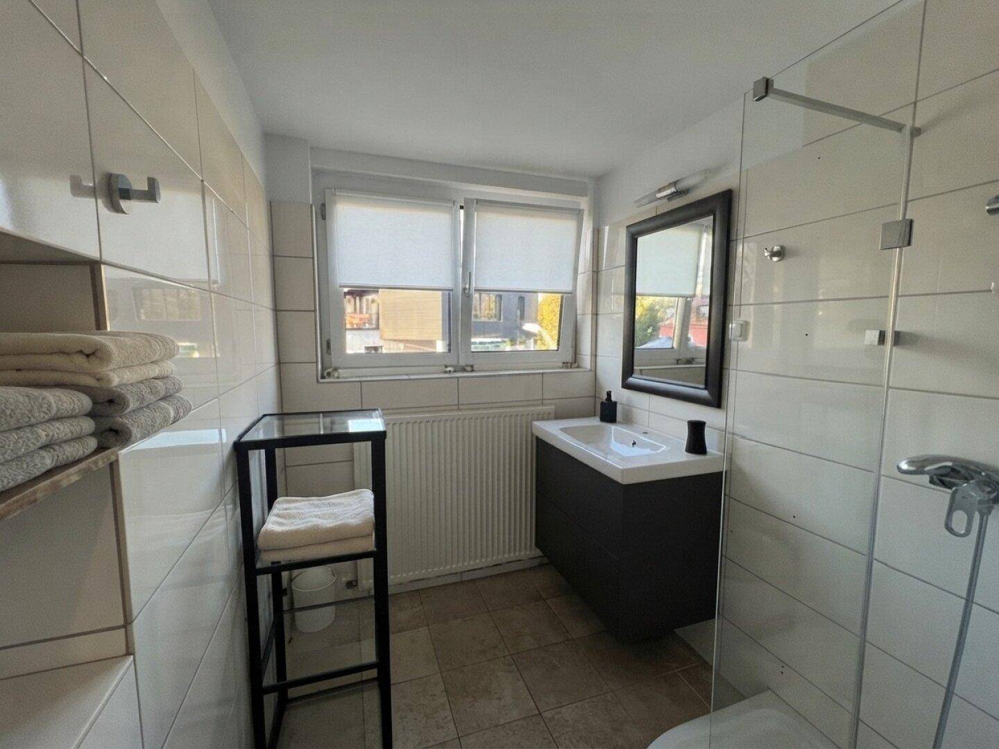 Modernes Badezimmer mit weißen Fliesen, Duschbereich, Einzelwaschbecken und Fenster für Tageslicht.