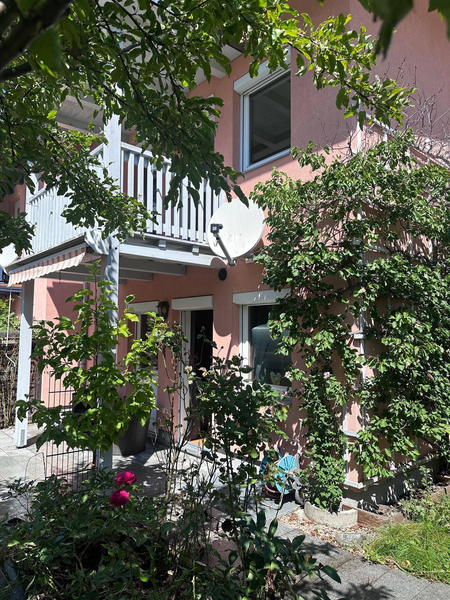 Rosa gestrichene Hausfassade mit üppiger Begrünung und einem Balkon im oberen Bereich.