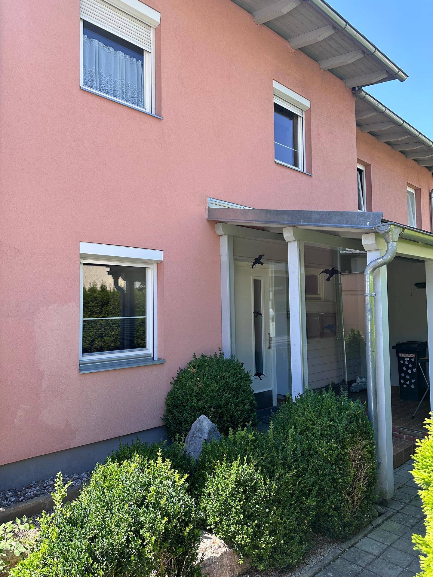 Vorderansicht des Hauses mit rosa Fassade, Fenstern und gepflegten Büschen im Vorgarten.