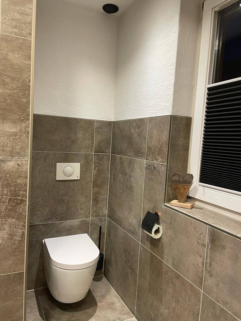 Modernes WC mit wandhängender Toilette, grauen Fliesen und einem Fenster für Tageslicht.
