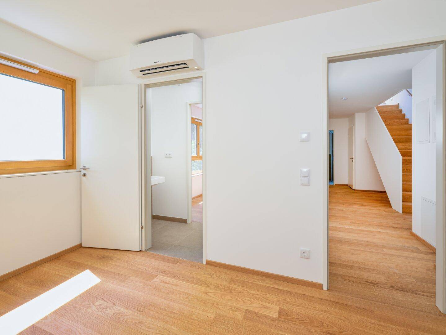 Helles Zimmer mit Holzboden, Klimaanlage und Zugang zu einem weiteren Raum oder Bad.