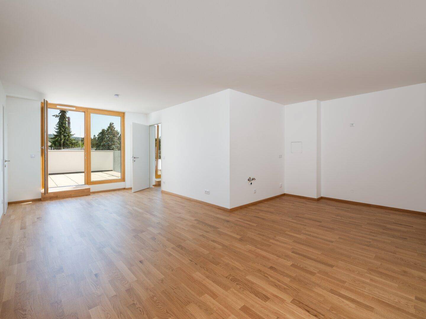 Heller, leerer Wohnraum mit hochwertigem Parkettboden und großen Fenstern mit Zugang zum Balkon.