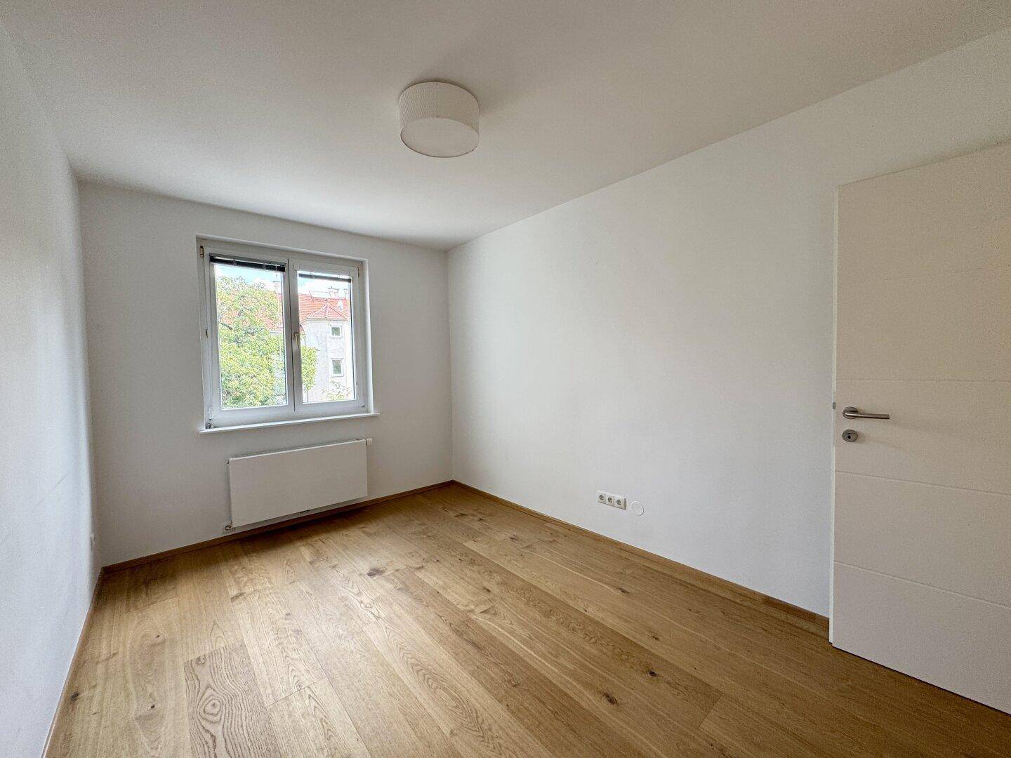 Helles Schlafzimmer mit Holzboden, weiß gestrichenen Wänden und Blick ins Grüne.