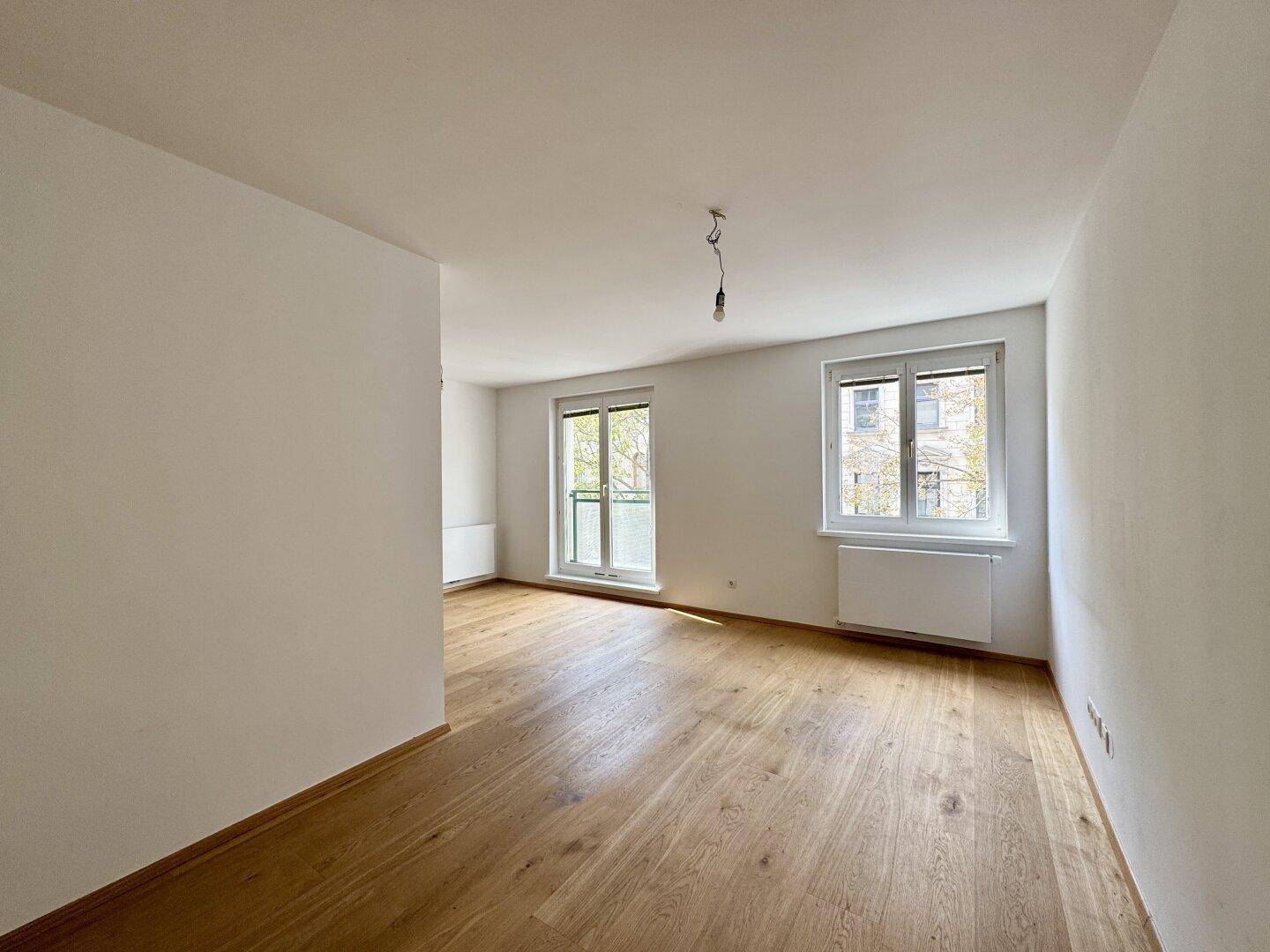 Heller Wohnraum mit Holzboden, großem Fenster und Zugang zu einem französischen Balkon.