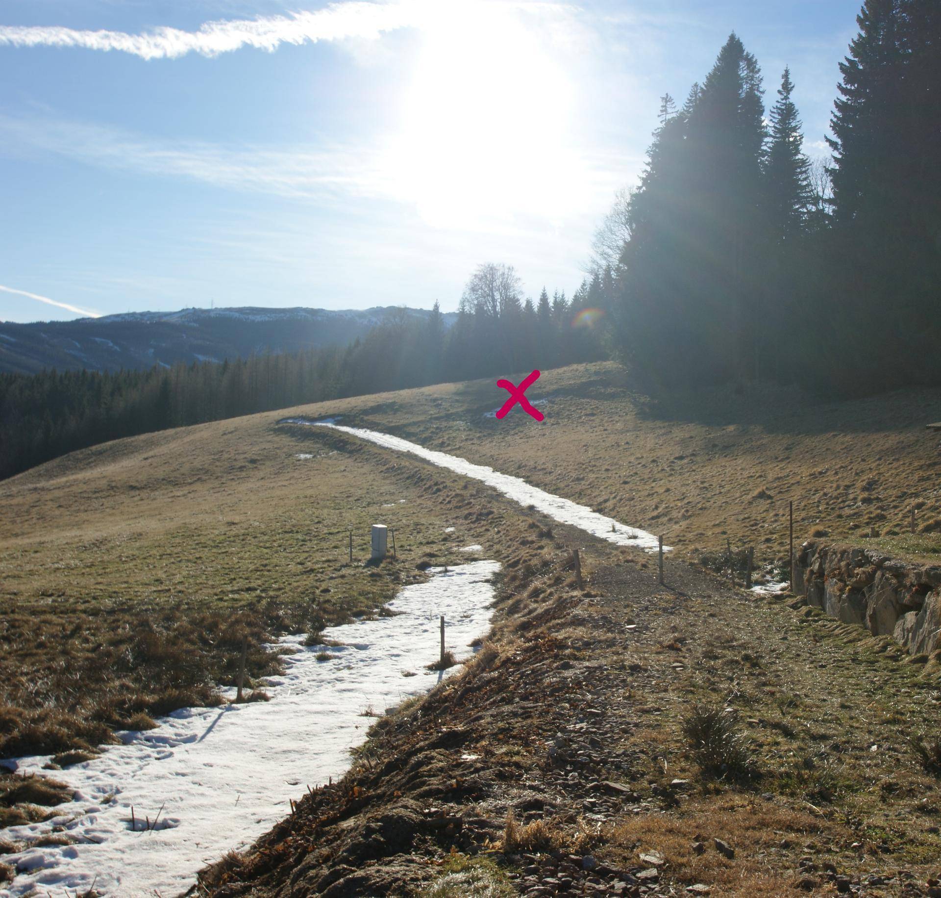 Winterliche Landschaft mit einem verschneiten Pfad, umgeben von Hügeln und Bäumen unter strahlendem Himmel.