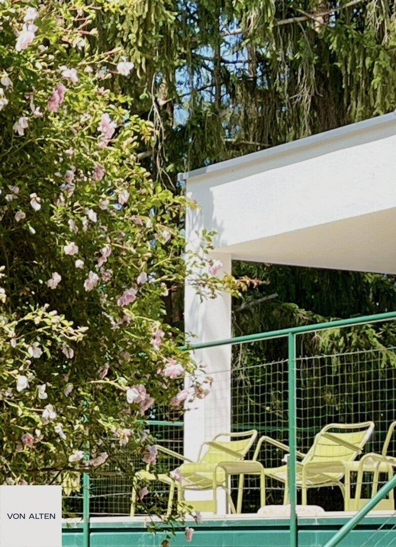 Teilansicht einer Terrasse mit blühenden Pflanzen und grünen Gartenmöbeln im Sonnenlicht.