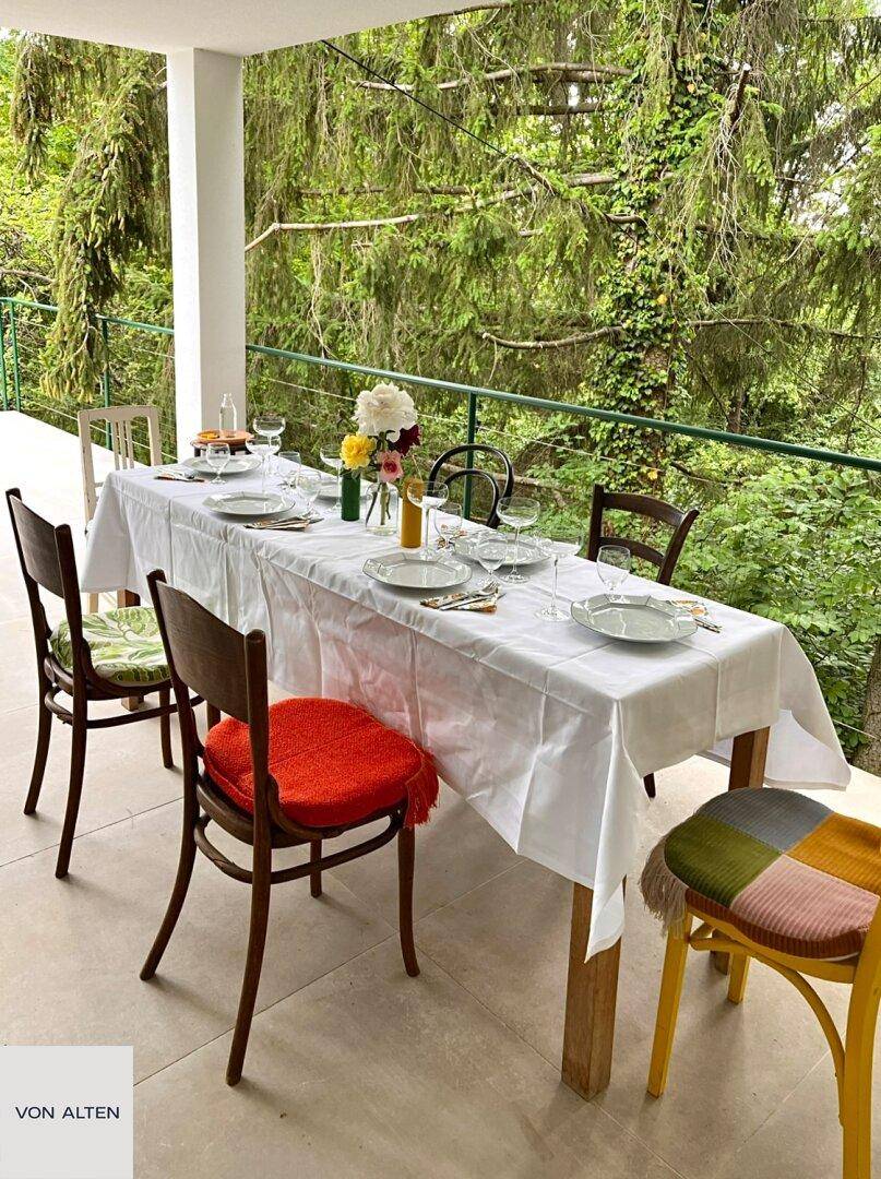 Überdachter Balkon mit gedecktem Esstisch und Stühlen, umgeben von üppigem Grün.
