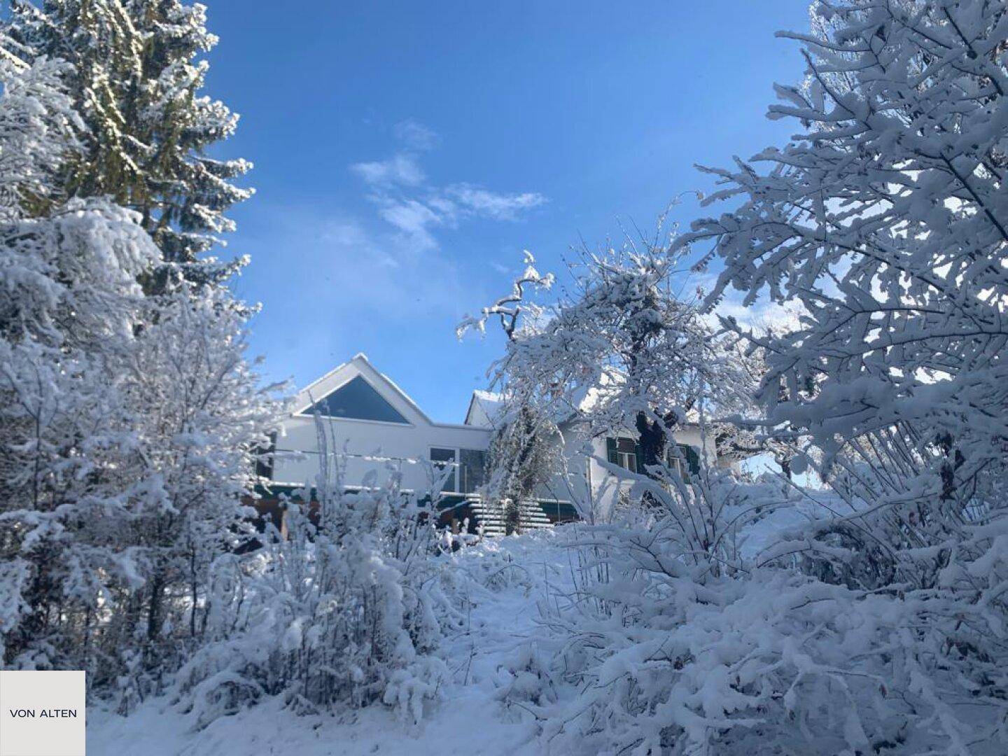 Haus in winterlicher Landschaft, umgeben von schneebedeckten Bäumen unter blauem Himmel.