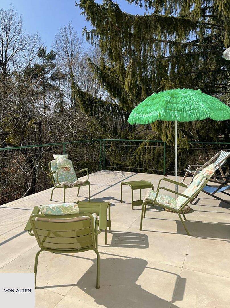 Sonnenterrasse mit grünen Gartenmöbeln und einem Sonnenschirm, umgeben von Bäumen.