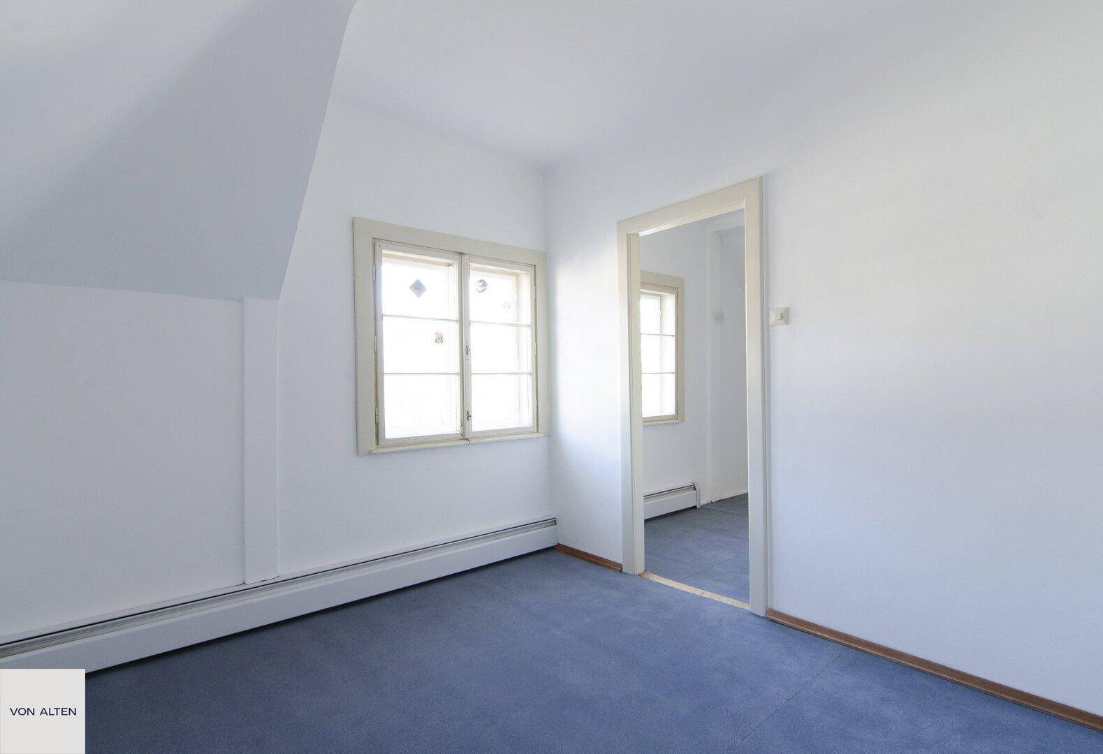 Dachgeschosszimmer mit blauem Teppichboden, schrägen Wänden und Fenster.