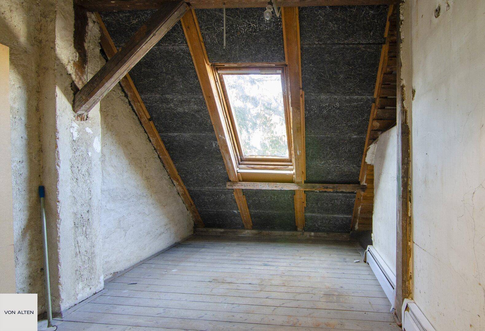Unausgebauter Dachbodenbereich mit sichtbaren Holzbalken, Dachfenster und Dielenboden.