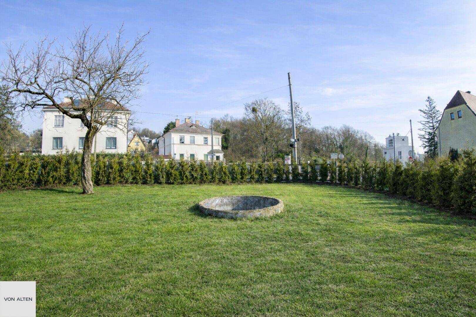 Großer, gepflegter Garten mit Rasenfläche, Hecke und einem runden Steinelement.