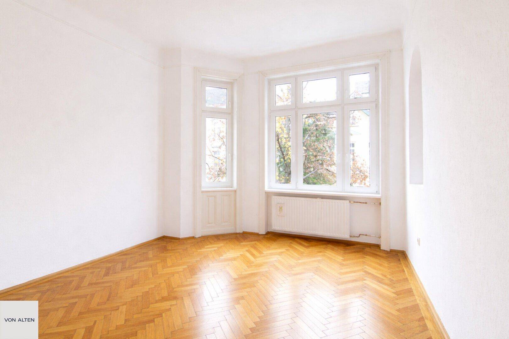 Großes, helles Zimmer mit Fischgrätparkett und einem markanten Erkerfenster, sehr einladend.