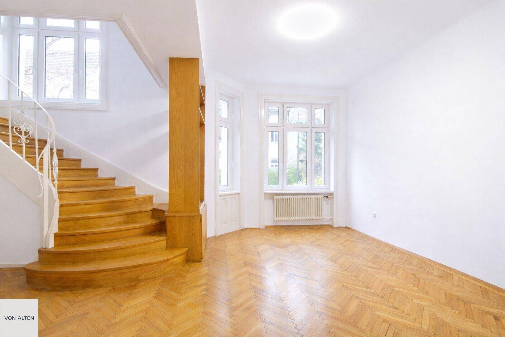 Heller Eingangsbereich mit geschwungener Holztreppe und Fischgrätparkettboden, gut beleuchtet.