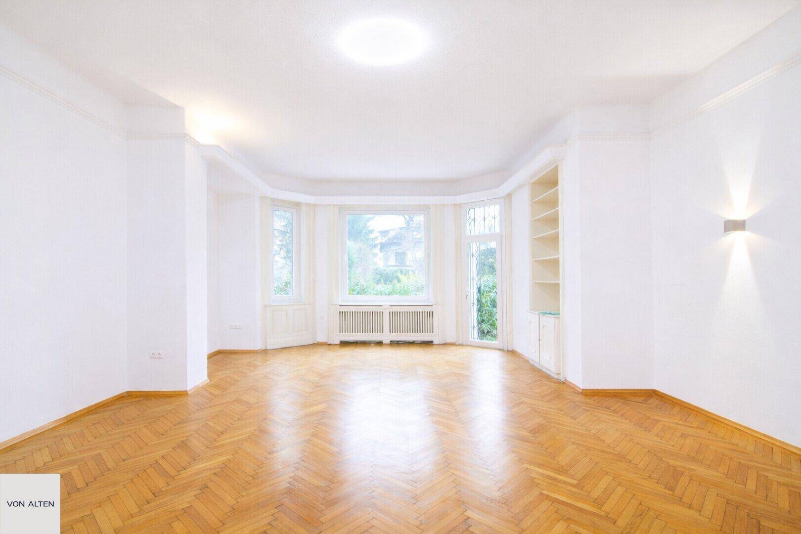Lichtdurchflutetes Wohnzimmer mit Erkerfenstern, Fischgrätparkett und praktischen Einbauregalen.