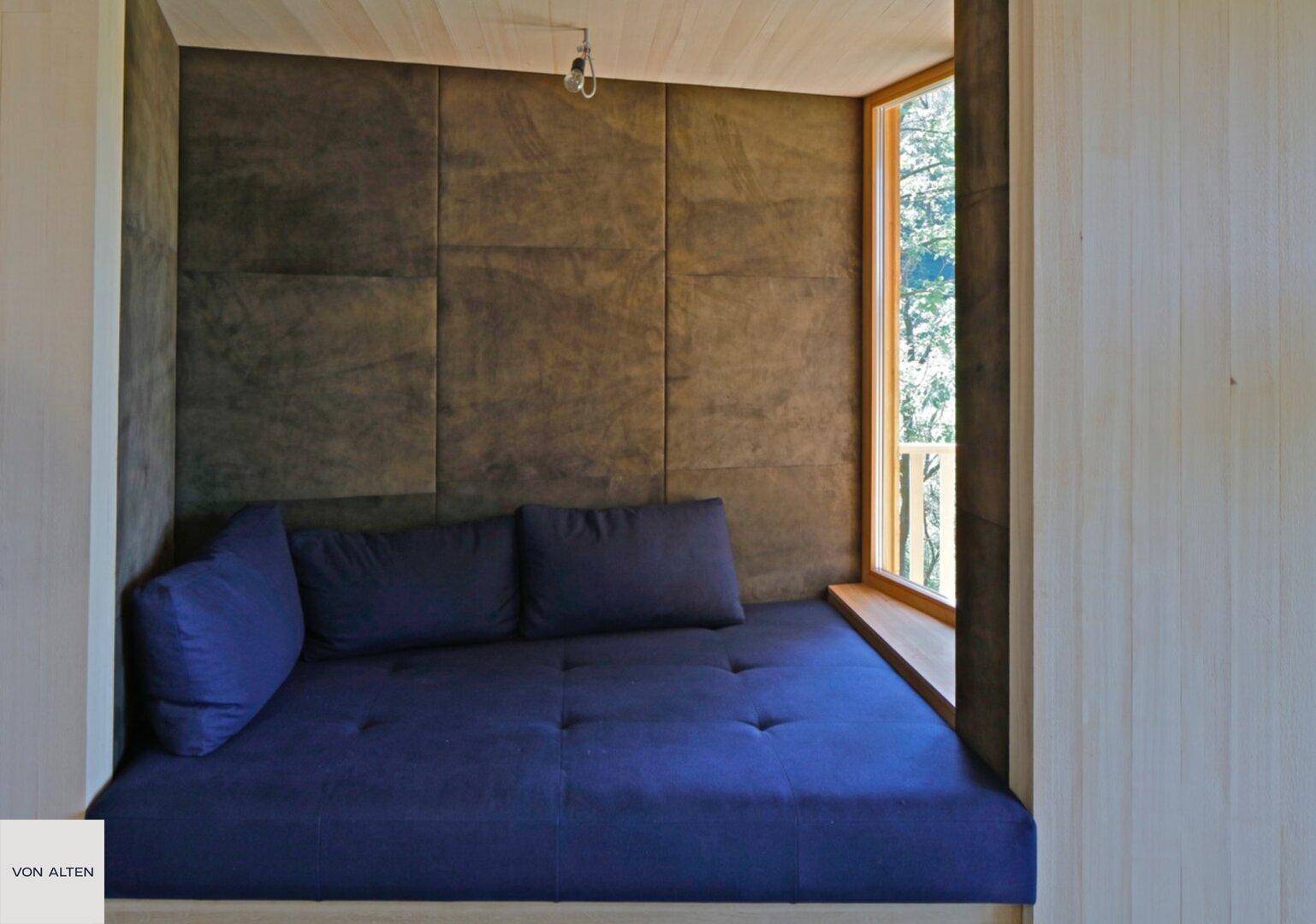 Gemütliche Schlafnische mit blauem Polsterbett und kleinem Fenster mit Waldblick.