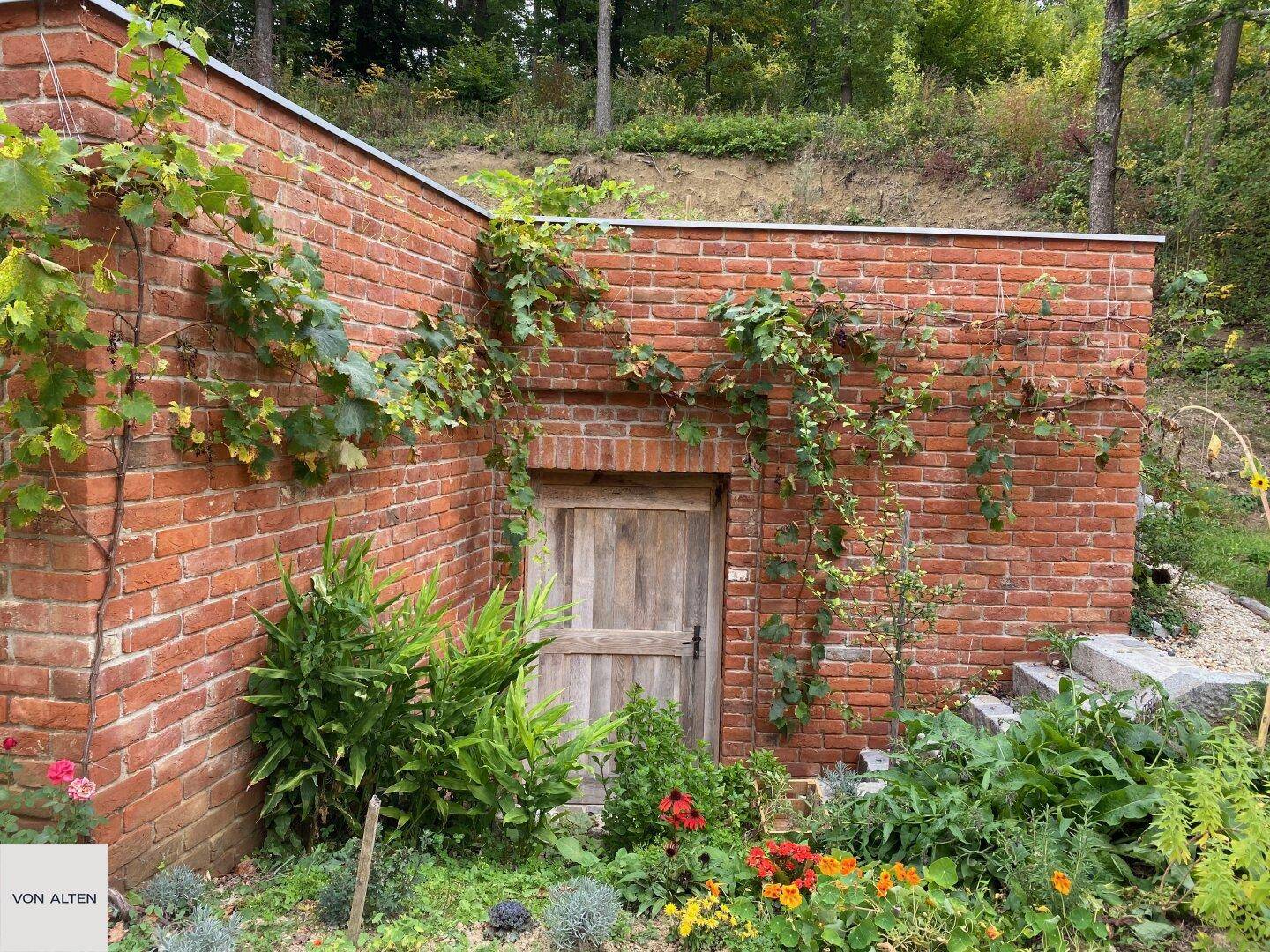 Alter Ziegelbau mit Holztür, umrankt von grünen Pflanzen und Blumenbeeten.